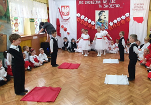 Dziewczynki w białych sukienkach z czerwonymi paskami oraz chłopcy ubrani na galowo wykonują układ taneczny z szarfami.